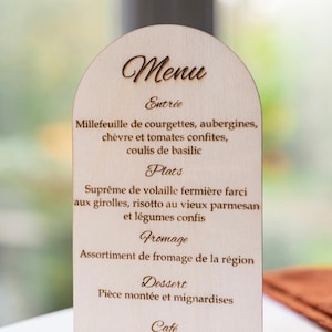 Peut inclure: Un menu en bois avec un sommet en forme d'arche arrondie. Le panneau est brun clair et a une bordure brun foncé. Le texte sur le panneau est en français et se lit comme suit : "Menu", "Entrée", "Millefeuille de courgettes, aubergines, chèvre et tomates confites, coulis de basilic", "Plats", "Suprême de volaille fermière farci aux girolles, risotto au vieux parmesan et légumes confis", "Fromage", "Assortiment de fromage de la région", "Dessert", "Pièce montée et mignardises", et "Café".