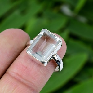 Puede incluir: Un anillo de plata con una piedra preciosa rectangular transparente. El anillo está en un dedo.