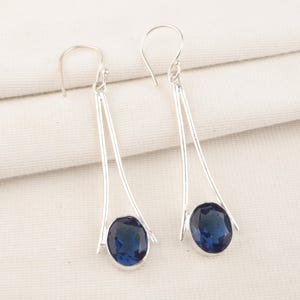 Pode incluir: Um par de brincos pendurados em prata. Cada brinco apresenta uma barra longa e fina de prata com uma pedra preciosa oval azul escuro na parte inferior. Os brincos penduram de um gancho simples.