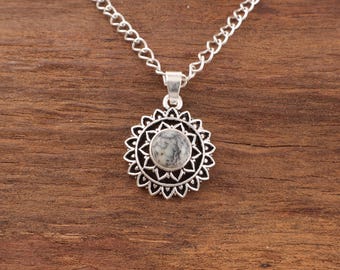 May include: A silver necklace with a pendant featuring a round, patterned stone set within a sunburst design. The chain is a delicate link style, and the pendant is centered. The background is a wooden surface.