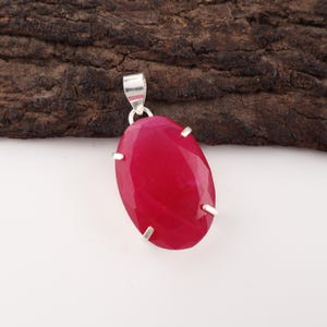 May include: A vivid red oval pendant with a silver bail and prongs. The gemstone is set against a dark wood background, creating a striking contrast. The pendant is a piece of jewellery.