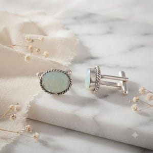 May include: A pair of silver cufflinks featuring oval-shaped, iridescent opal-like stones set within a twisted rope-style metal border. The cufflinks have a polished silver finish and a standard T-bar toggle mechanism for secure fastening.