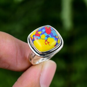 May include: A silver ring with a square-shaped yellow, blue, and red gemstone. The gemstone has a marbled pattern.