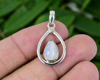 May include: A silver pendant with a teardrop-shaped moonstone. The moonstone has a pearlescent white color with blue and gray hues. The pendant is suspended from a silver bail and is held in a hand.