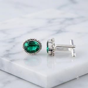 May include: A pair of silver cufflinks featuring oval-shaped, faceted emerald green stones. Each cufflink has a twisted rope-style border surrounding the central green gem, with a standard T-bar toggle mechanism attached to the back of the setting.