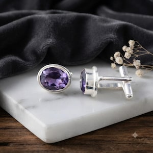 May include: A pair of silver cufflinks featuring oval-cut purple amethyst gemstones. The cufflinks have a polished metallic finish and a classic T-bar toggle mechanism, showcasing the deep violet facets of the stones in a clear, bright setting.