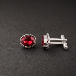 Puede incluir: Un par de gemelos plateados con piedras facetadas de color rojo intenso en forma ovalada. Cada gemelo está enmarcado por un borde de metal trenzado y cuenta con un mecanismo giratorio en forma de T para asegurar el accesorio al puño.