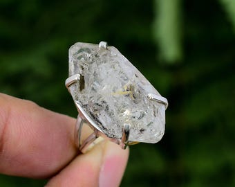 May include: A silver ring with a large, clear, faceted quartz crystal set in a bezel setting. The crystal has a rough, natural texture and is set in a simple, modern band.