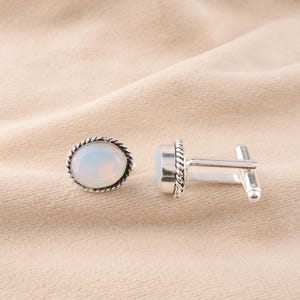 May include: A pair of silver cufflinks with a white opal stone in each. The cufflinks have a rope-like design around the edge of the stone.