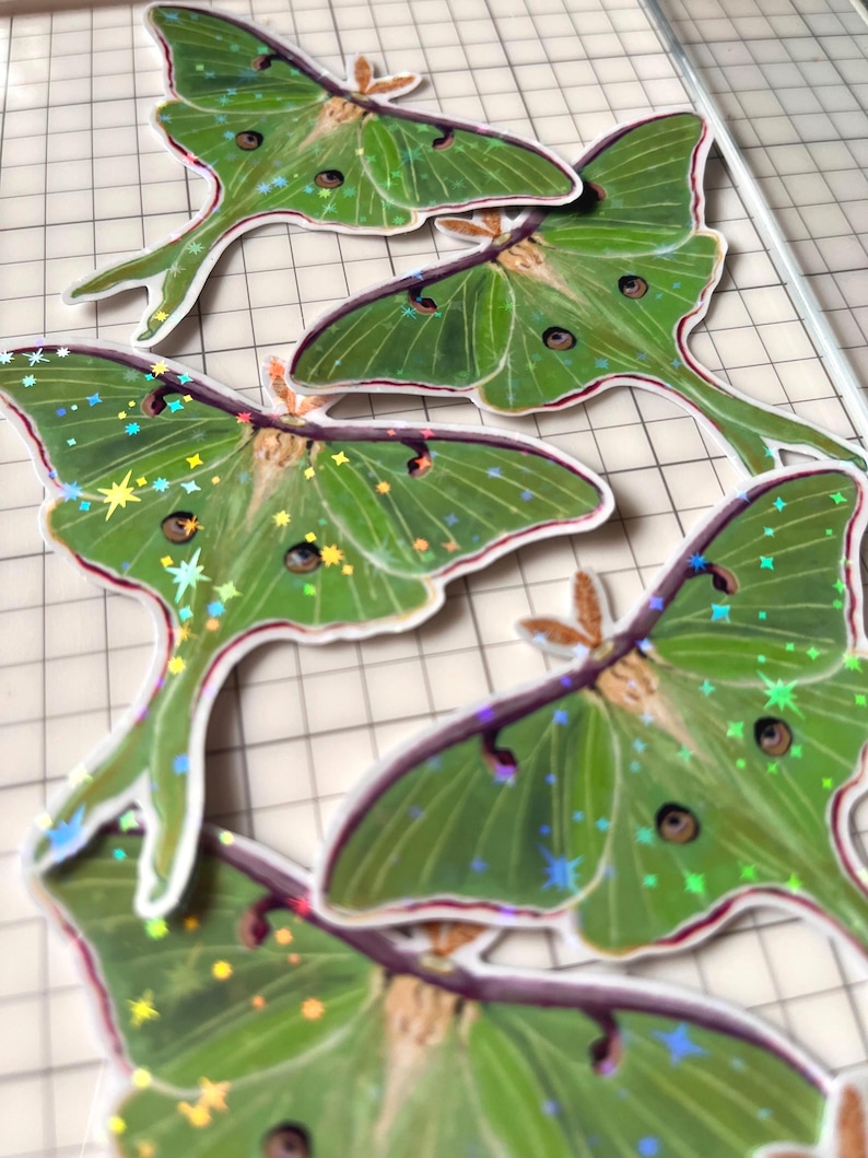 May include: A collection of die-cut stickers featuring green Luna moths with long, trailing hindwings. Each moth displays a detailed wing pattern with dark eyespots and a holographic finish that reflects light as small, colourful star-shaped sparkles.