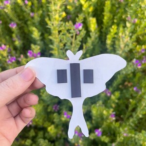 May include: A white moth-shaped cutout featuring three rectangular black magnetic strips attached to the back. The moth has a wide wing span and a long, slender tail section, with the magnets positioned in the centre of the body area.