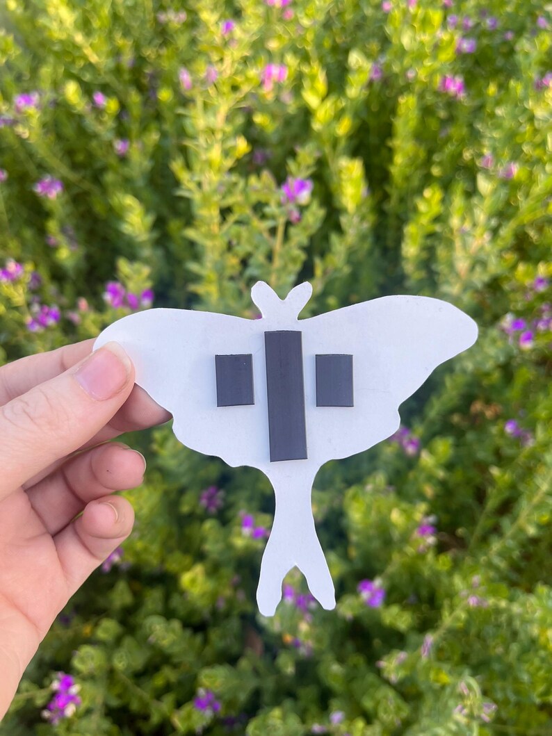 May include: A white moth-shaped cutout featuring three rectangular black magnetic strips attached to the back. The moth has a wide wing span and a long, slender tail section, with the magnets positioned in the centre of the body area.