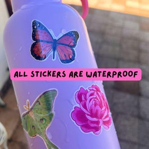 May include: A light purple water bottle decorated with three stickers: a pink and black butterfly, a green moth, and a bright pink flower. A pink horizontal banner across the center displays the black text: ALL STICKERS ARE WATERPROOF.