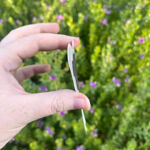 May include: A thin, flat, light-coloured object with a dark, rectangular magnetic strip attached to its side. The item is shown from a side profile view, revealing its slim construction and the dark magnetic component secured to the surface.