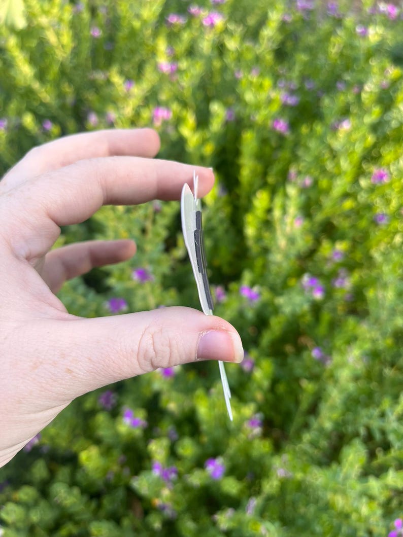 May include: A thin, flat, light-coloured object with a dark, rectangular magnetic strip attached to its side. The item is shown from a side profile view, revealing its slim construction and the dark magnetic component secured to the surface.