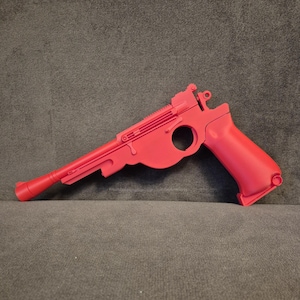 Può includere: Una pistola giocattolo rosso brillante con una canna lunga e un'impugnatura curva. La pistola giocattolo ha un'apertura circolare e creste dettagliate sulla parte superiore. Il giocattolo è realizzato in un materiale solido e poggia su una superficie grigia.