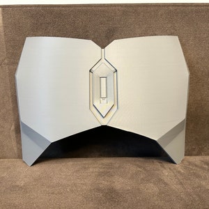 Pu&ograve; includere: Un pezzo toracico in plastica grigia stampato in 3D con un design geometrico. Il pezzo toracico ha un ritaglio al centro.