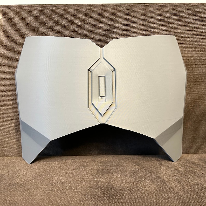 Pu&ograve; includere: Un pezzo toracico in plastica grigia stampato in 3D con un design geometrico. Il pezzo toracico ha un ritaglio al centro.