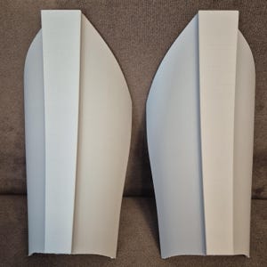 Op de afbeelding: Twee lichtgrijze armbeschermers, waarschijnlijk voor kostuum of cosplay. Elk heeft een centrale, verhoogde verticale sectie en een afgeronde, gladde buitenrand. De items worden getoond op een bruin stoffen oppervlak.