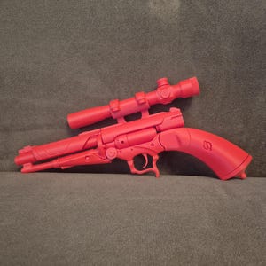 Puede incluir: Una pistola de juguete de color rojo brillante con mira telescópica, sobre una superficie gris. La pistola tiene gatillo, cañón y empuñadura, con la mira montada en la parte superior. El diseño general sugiere un tema futurista o de ciencia ficción.