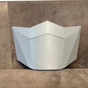 Pu&ograve; includere: Una maschera bianca di forma geometrica stampata in 3D con una parte superiore a punta e una parte inferiore arrotondata.