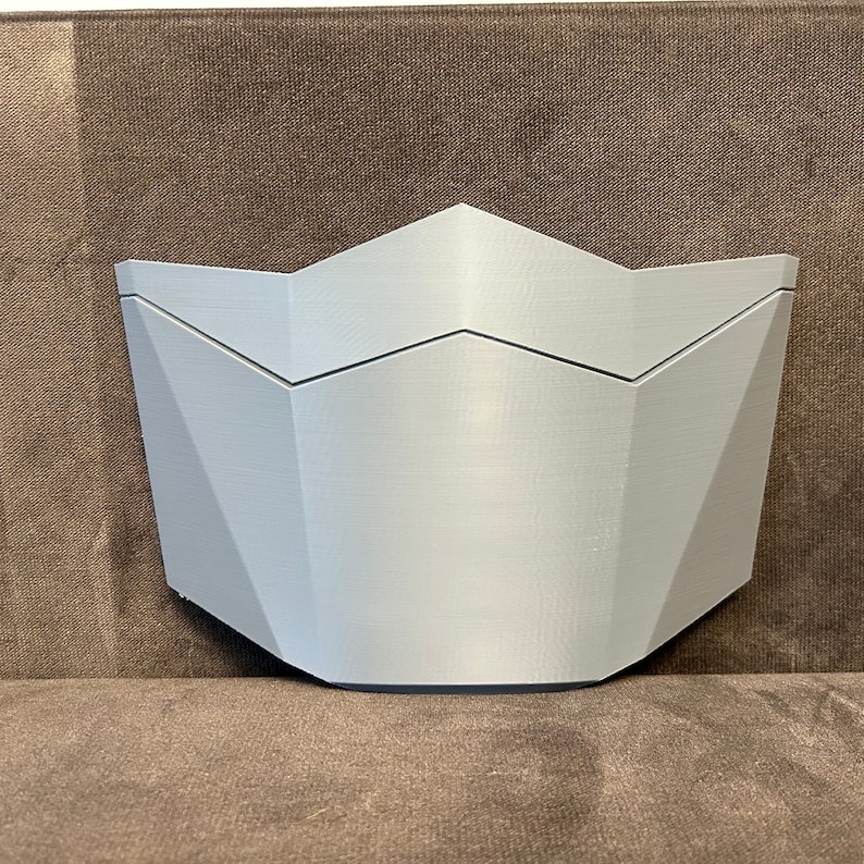 Pu&ograve; includere: Una maschera bianca di forma geometrica stampata in 3D con una parte superiore a punta e una parte inferiore arrotondata.