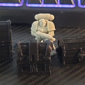 Puede incluir: Tres peque&ntilde;os cofres de madera negra con detalles intrincados. Los cofres est&aacute;n cerrados y tienen detalles met&aacute;licos. Una figura en miniatura de pl&aacute;stico gris de un marine espacial se encuentra frente a los cofres.