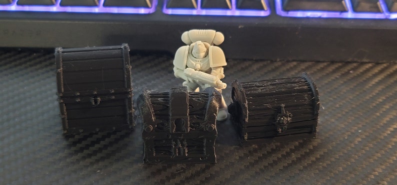 Puede incluir: Tres peque&ntilde;os cofres de madera negra con detalles intrincados. Los cofres est&aacute;n cerrados y tienen detalles met&aacute;licos. Una figura en miniatura de pl&aacute;stico gris de un marine espacial se encuentra frente a los cofres.