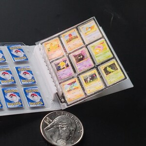 May include: A miniature trading card binder containing small cards with blue and white backs featuring the Pok&eacute;mon logo, and front-facing cards with various colorful character illustrations. A silver United States quarter coin is placed nearby.