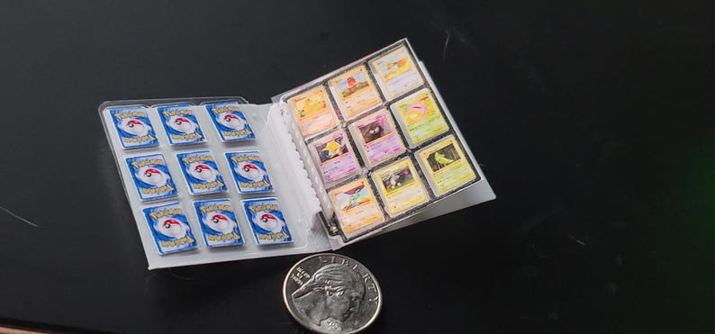 May include: A miniature trading card binder containing small cards with blue and white backs featuring the Pok&eacute;mon logo, and front-facing cards with various colorful character illustrations. A silver United States quarter coin is placed nearby.