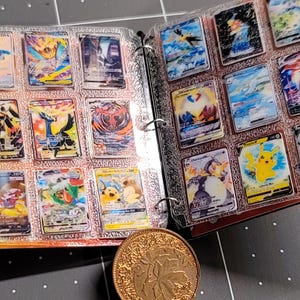 May include: A yellow binder with a repeating red and white pokeball pattern, containing clear plastic sleeves filled with various colorful trading cards featuring illustrated creatures. A round, textured gold-colored metal coin sits in front.