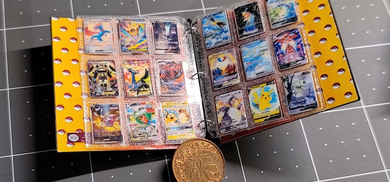 May include: A yellow binder with a repeating red and white pokeball pattern, containing clear plastic sleeves filled with various colorful trading cards featuring illustrated creatures. A round, textured gold-colored metal coin sits in front.