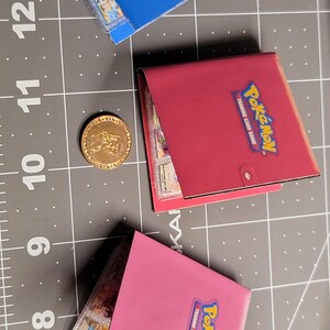 May include: Three small square Pok&eacute;mon Trading Card Game binders in blue, red, and pink, each featuring the Pok&eacute;mon logo on the front cover. A single gold-colored circular coin is positioned between the blue and red binders on a cutting mat.