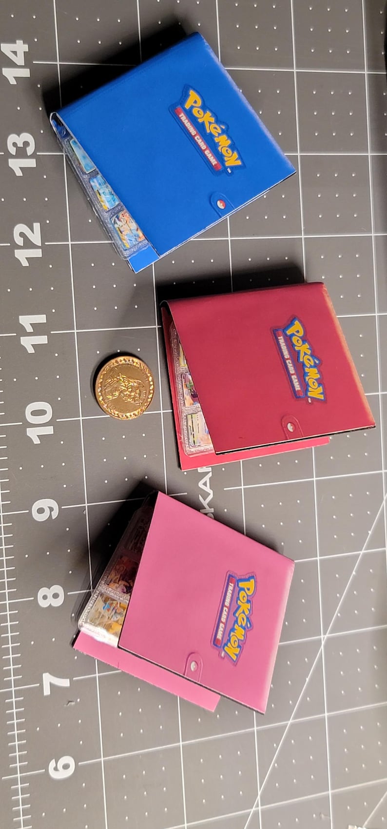 May include: Three small square Pok&eacute;mon Trading Card Game binders in blue, red, and pink, each featuring the Pok&eacute;mon logo on the front cover. A single gold-colored circular coin is positioned between the blue and red binders on a cutting mat.