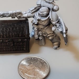 Puede incluir: Dos peque&ntilde;os cofres del tesoro de madera con detalles intrincados, pintados en un color marr&oacute;n oscuro. Una figura miniatura de Space Marine de pl&aacute;stico gris se encuentra junto a los cofres, sosteniendo un arma. Una moneda de euro espa&ntilde;ola se incluye para la escala.