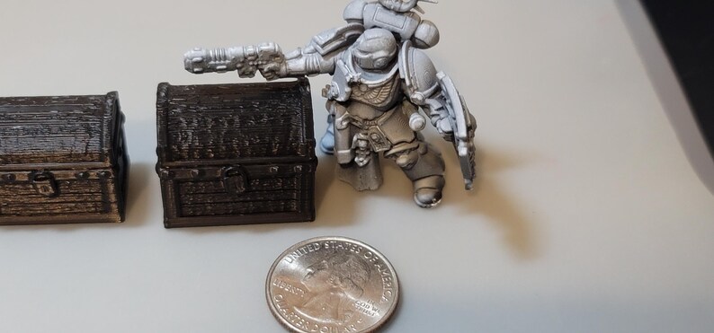 Puede incluir: Dos peque&ntilde;os cofres del tesoro de madera con detalles intrincados, pintados en un color marr&oacute;n oscuro. Una figura miniatura de Space Marine de pl&aacute;stico gris se encuentra junto a los cofres, sosteniendo un arma. Una moneda de euro espa&ntilde;ola se incluye para la escala.