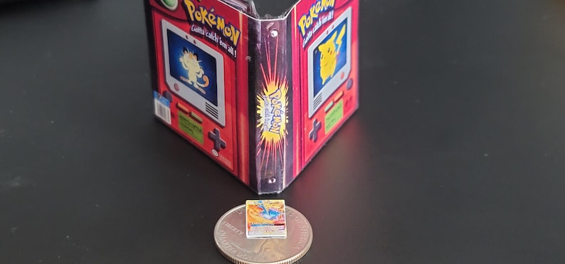 May include: A miniature Pok&eacute;mon-themed binder featuring red and black graphics with images of Meowth and Pikachu on the front panels. A tiny, rectangular Pok&eacute;mon trading card rests on top of a United States quarter coin for scale comparison.