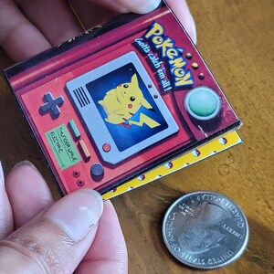 May include: A square pin featuring a red handheld gaming console graphic with a yellow Pikachu character on the screen, the text Pok&eacute;mon gotta catch 'em all!, and a green button. A United States quarter is placed next to the pin for scale.