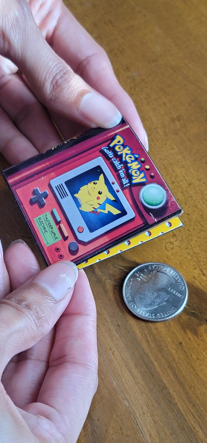 May include: A square pin featuring a red handheld gaming console graphic with a yellow Pikachu character on the screen, the text Pok&eacute;mon gotta catch 'em all!, and a green button. A United States quarter is placed next to the pin for scale.