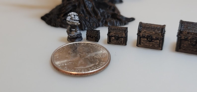 Puede incluir: Una colecci&oacute;n de cofres del tesoro en miniatura, algunos con detalles intrincados, y una peque&ntilde;a figura gris detallada. Los cofres son de varios tama&ntilde;os y est&aacute;n hechos de un material oscuro y texturizado. Se incluye una moneda de 25 centavos de d&oacute;lar estadounidense para la escala.