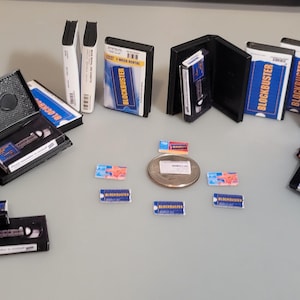 May include: A collection of miniature VHS tapes, some with the Blockbuster logo, and a few empty cases. There are also several small blue and red rectangular stickers, and a silver coin.