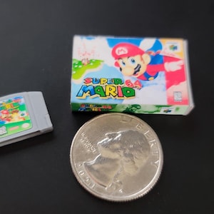 May include: A miniature Super Mario 64 video game cartridge and box set next to a quarter. The game box features Mario in a red hat and blue overalls. The cartridge is gray with a colorful label.