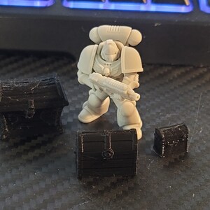 Puede incluir: Una miniatura de pl&aacute;stico blanco de un marine espacial sosteniendo un arma, de pie frente a tres cofres del tesoro de pl&aacute;stico negro.