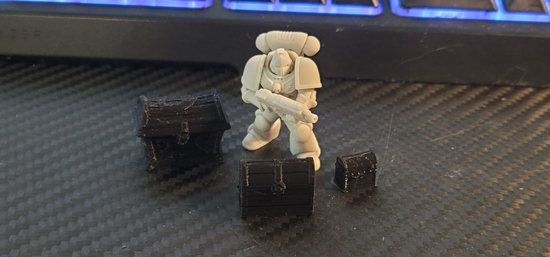 Puede incluir: Una miniatura de pl&aacute;stico blanco de un marine espacial sosteniendo un arma, de pie frente a tres cofres del tesoro de pl&aacute;stico negro.