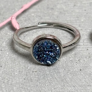 Puede incluir: Un anillo plateado con una piedra druzy azul redonda engastada en bisel. El anillo es ajustable. La piedra brilla. El anillo se muestra sobre una superficie gris con un tallo rosa en el fondo.