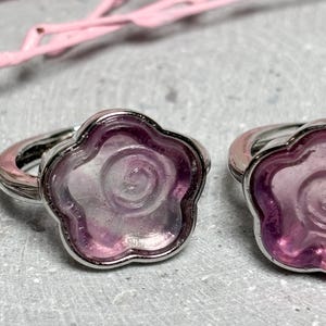 Könnte beinhalten: Zwei ringförmige Schmuckstücke mit einem blütenförmigen Design. Sie haben ein silbernes Band und einen durchscheinenden, hellvioletten Mittelteil. Die Blütenblätter sind dunkelviolett umrandet. Die Ringe sind vor einem grauen Hintergrund platziert.