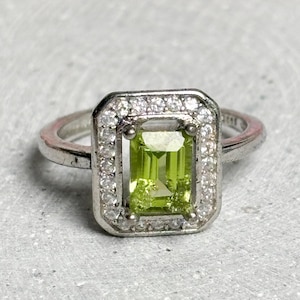 Peut inclure: Une bague en argent ornée d'une pierre précieuse rectangulaire vert clair, entourée d'un halo de petites pierres claires. Le corps de la bague est également en argent, créant un design classique et élégant. La pierre verte est le point central.