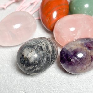 May include: Assortment of polished gemstone eggs in various colors, including pink, red, green, gray, and purple. The eggs are smooth and glossy, with natural patterns and variations in color. The gemstones are arranged on a white surface.