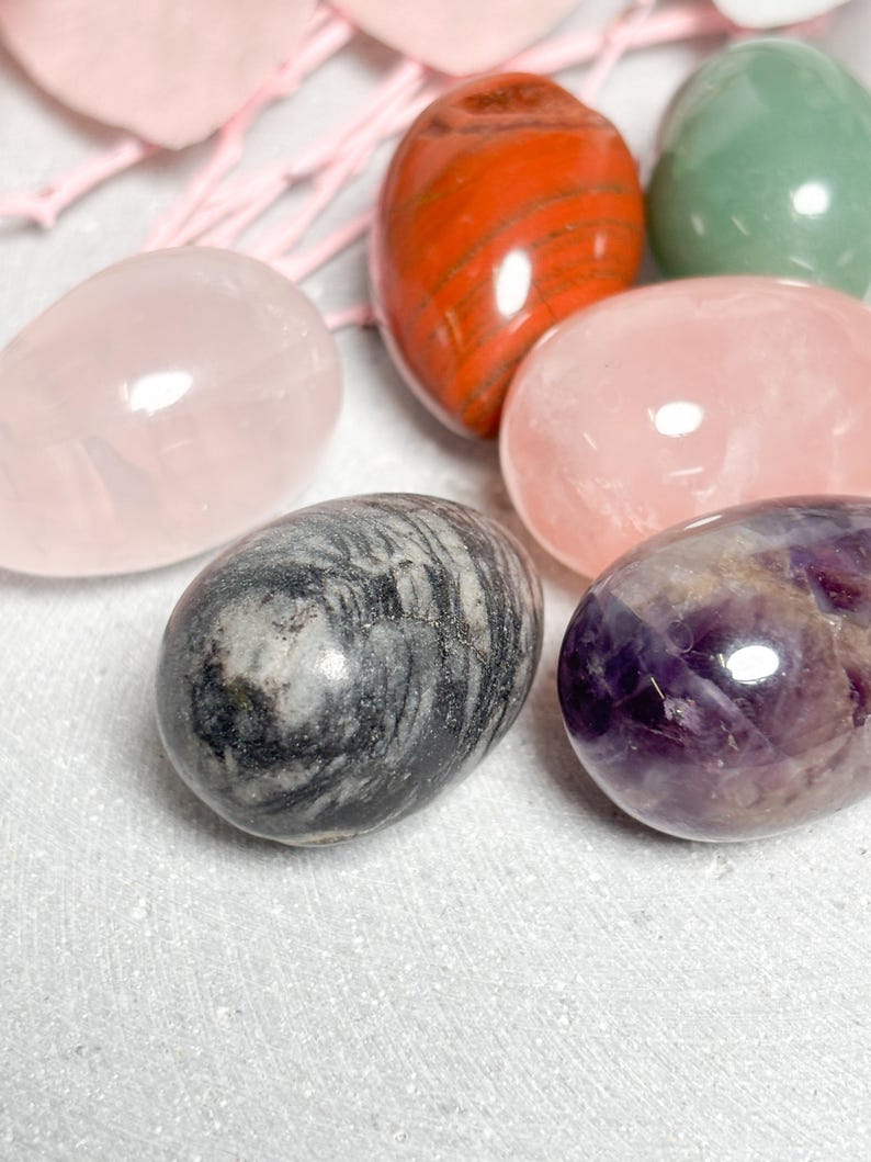 May include: Assortment of polished gemstone eggs in various colors, including pink, red, green, gray, and purple. The eggs are smooth and glossy, with natural patterns and variations in color. The gemstones are arranged on a white surface.