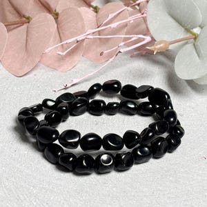 Peut inclure: Deux bracelets de perles noires. Les perles sont lisses et de forme ovale, avec une finition brillante. Les bracelets sont empilés, créant un effet superposé. Le fond comprend des feuilles décoratives roses et blanches.